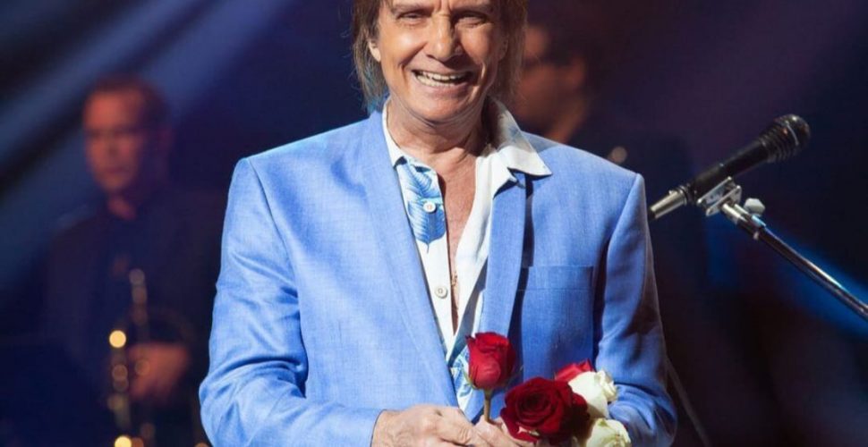 Roberto Carlos em um show com terno azul segurando uma rosa vermelha.