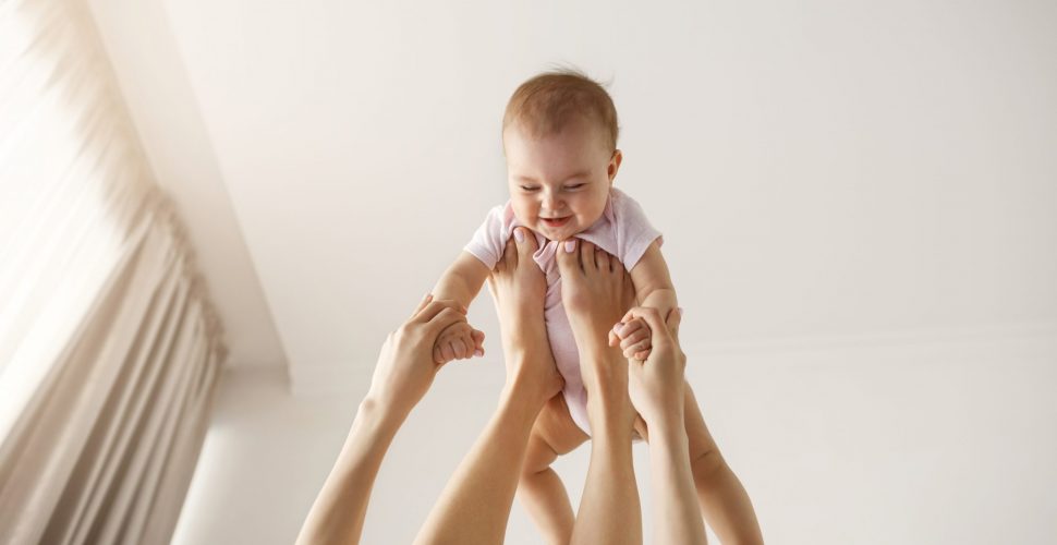 Young cheerful mother playing with her newborn baby lying on bed at home. Copy space.